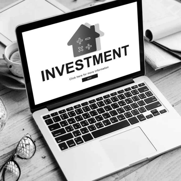 A laptop screen displaying the word "INVESTMENT" with a house icon, suggesting exploration of alternative investments, surrounded by a desk with notepads and glasses.
