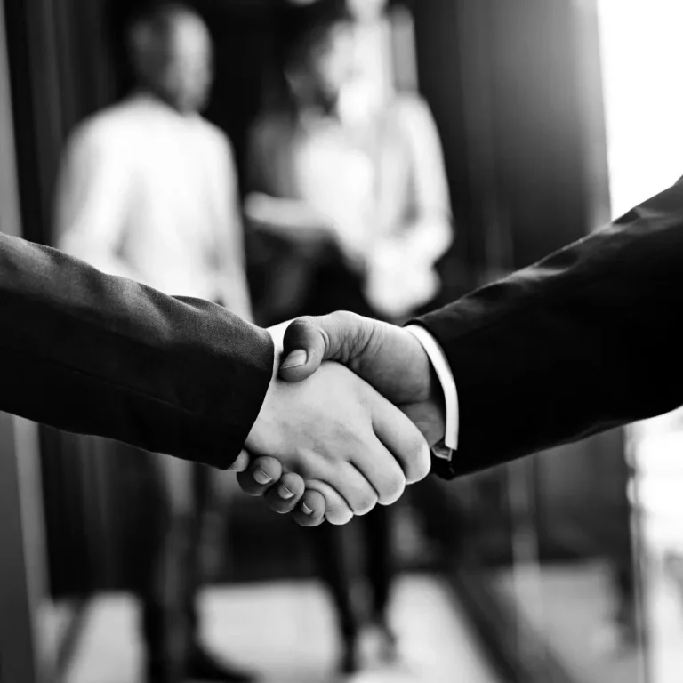 Two people in suits shake hands in a buy-sell agreement, with two blurred figures conversing in the background.