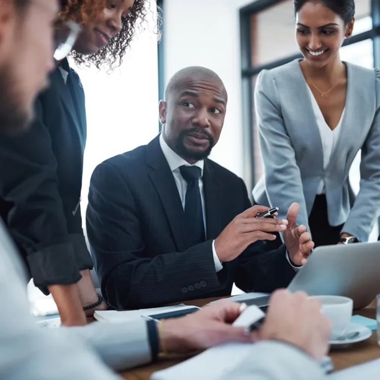 An independent financial advisor in a suit engages with a team around a conference table, discussing strategies over a laptop.