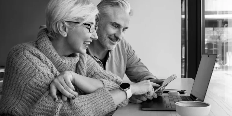 An older couple happily reviews tax reduction strategies for retirement on a laptop and smartphone at a table with a cup of coffee.