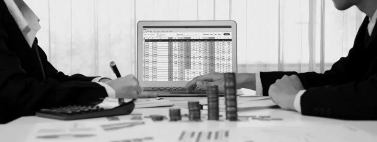 Two people in suits analyze financial data on a laptop, surrounded by coins and paperwork, highlighting the evaluation of tax loss harvesting worth.