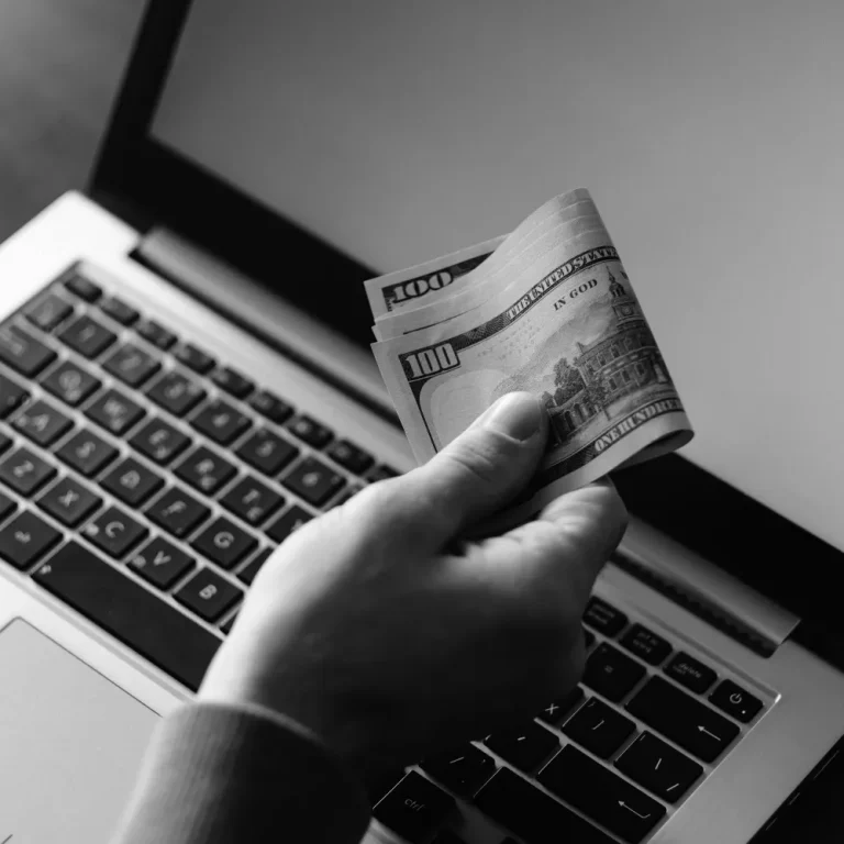 A person holds a stack of hundred-dollar bills over a laptop keyboard, symbolizing the concept of S Corp self-employment tax.