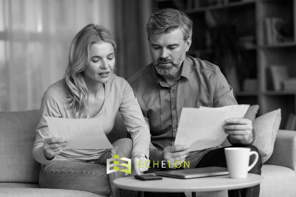 A couple sits on a couch reviewing beneficiary designation documents, with papers in hand and a coffee cup on the table.