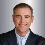 Smiling man in a dark suit and light shirt against a gradient background.