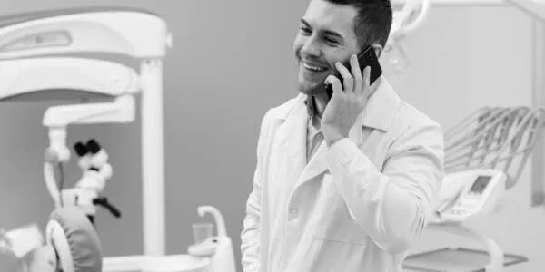 A man in a dental office, wearing a lab coat, smiles while talking on a cellphone.