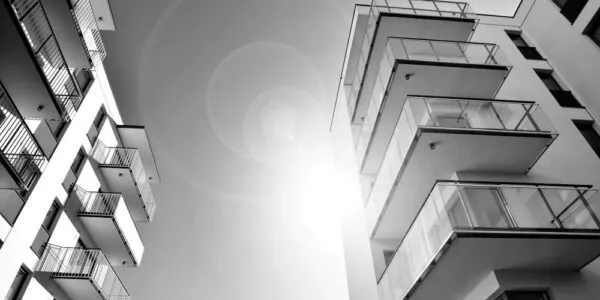 Two modern apartment buildings with glass balconies rise against a bright sun, creating lens flares in a black and white composition.