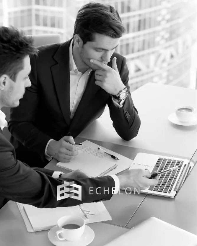 Two businessmen in suits are focused on a laptop, analyzing documents for business exit planning, with coffee cups and notebooks on the table.