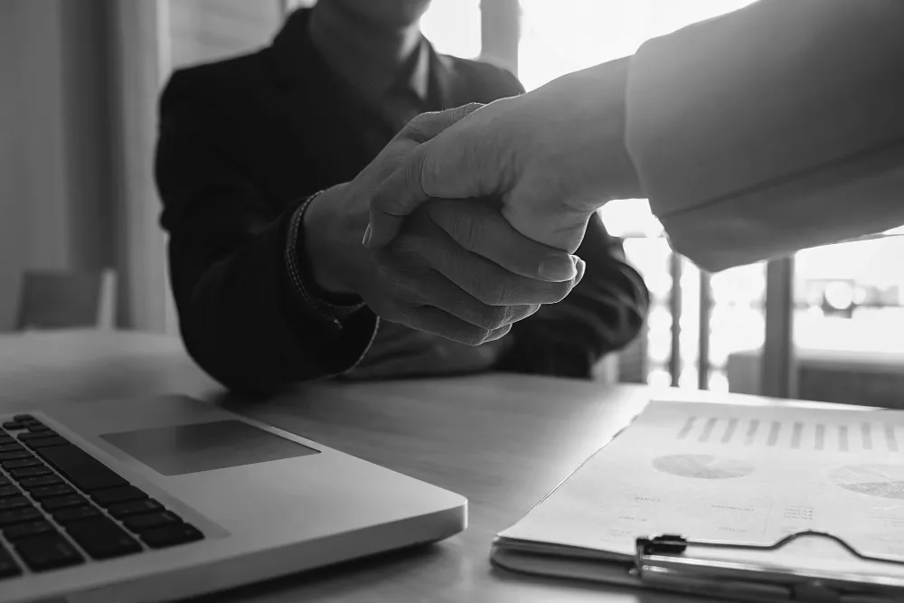 Two people shaking hands over a table with documents and a laptop, symbolizing a family limited partnership agreement.