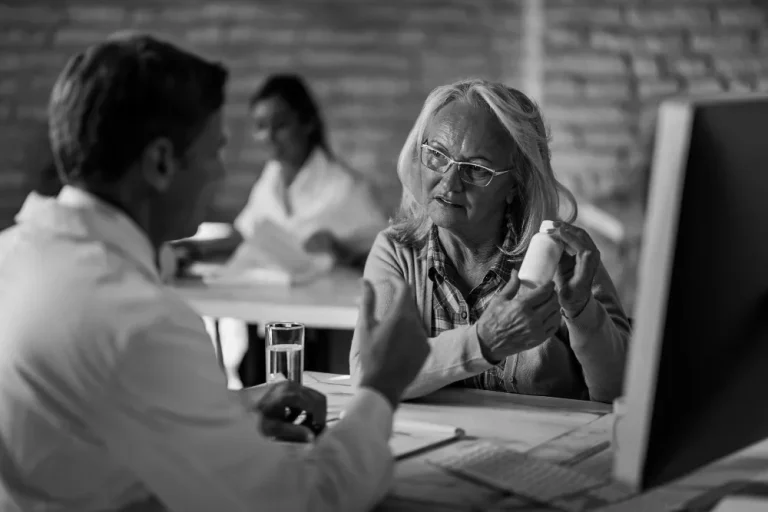 An older woman, identified as Irmaa, attentively discusses a medication bottle with a doctor in an office setting, with another medical professional in the background.