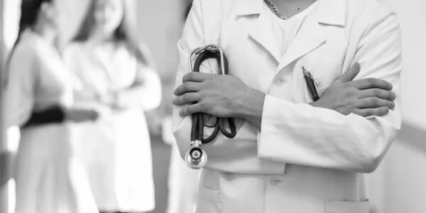 A doctor in a white coat stands with crossed arms holding a stethoscope, with two blurred figures in the background.