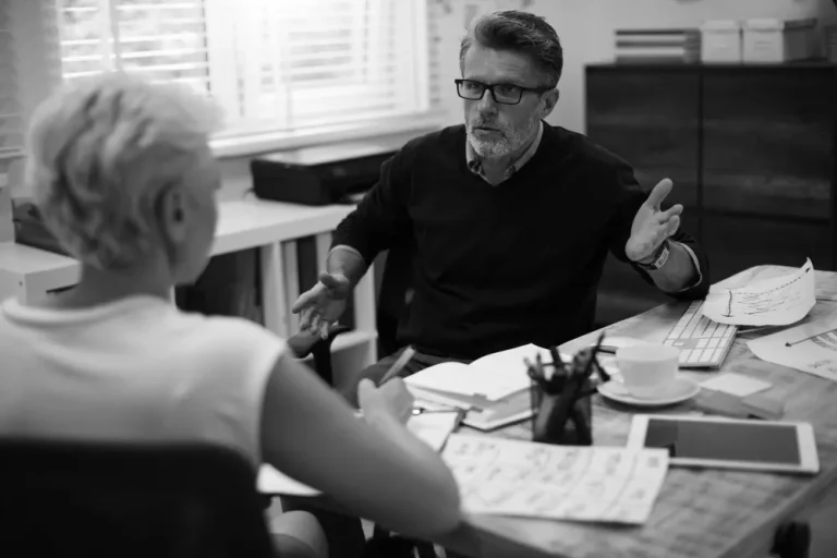 A man discusses retirement income planning with a woman at a cluttered desk, both seeming engaged and focused.