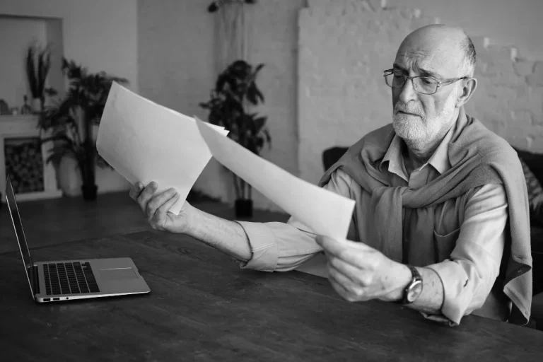 An elderly man, seated at a table with a laptop, intently examines documents related to a 401k rollover, wearing glasses and a sweater draped over his shoulders.