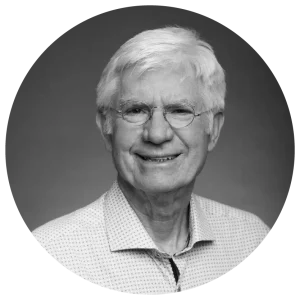 Elderly man with white hair and glasses smiling, wearing a light patterned shirt, set against a plain background.