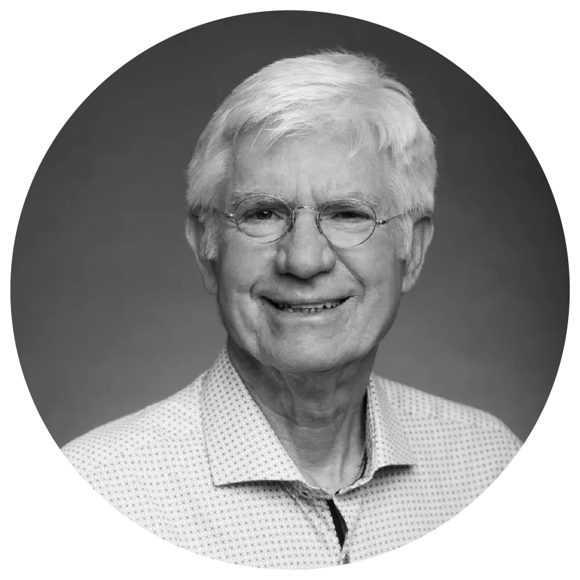 Elderly man with white hair and glasses smiling, wearing a light patterned shirt, set against a plain background.