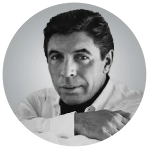 A grayscale portrait of a person with short dark hair and light skin, resting their arms on a surface, looking directly at the camera.