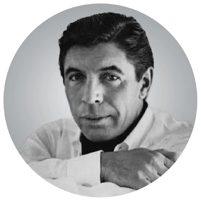 A grayscale portrait of a person with short dark hair and light skin, resting their arms on a surface, looking directly at the camera.